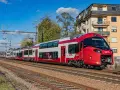 Nowoczesne, czerwono-białe piętrowe pociągi CFL 2410 pędzą po torach, mijając budynki i drzewa.