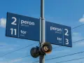 Niebieska tablica informacyjna na dworcu kolejowym wskazuje peron 2, tor 11 oraz peron 2, tor 3. W tle błękitne niebo z chmurami i linie trakcyjne.