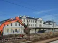 Koparka pracuje przy modernizacji dworca Skarżysko Kamienna. Widać budynek dworca, tory i pracowników w kamizelkach.