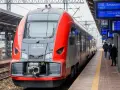 Czerwono-szary pociąg stoi na peronie, obok tablic informacyjnych z rozkładem jazdy.
