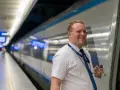 Uśmiechnięty konduktor PKP Intercity w białej koszuli i niebieskim krawacie stoi na peronie obok pociągu.