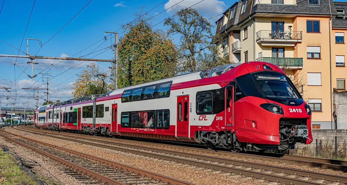 Nowoczesne, czerwono-białe piętrowe pociągi CFL 2410 pędzą po torach, mijając budynki i drzewa.
