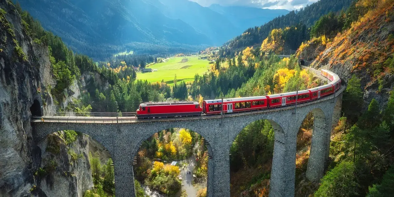 Czerwony pociąg Bernina Express przejeżdża przez kamienny wiadukt w górach jesienią.