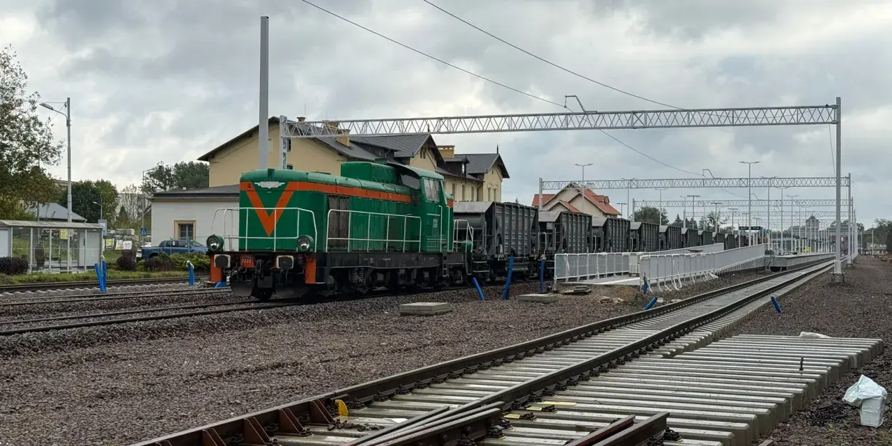 Zielona lokomotywa z pomarańczowymi pasami na torach. W tle budynki dworca i wagonów towarowych. Sprawdzam kętrzyn-olsztyn rozkład jazdy.