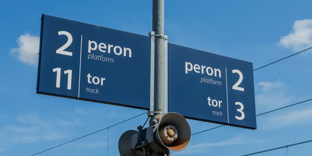 Niebieska tablica informacyjna na dworcu kolejowym wskazuje peron 2, tor 11 oraz peron 2, tor 3. W tle błękitne niebo z chmurami i linie trakcyjne.