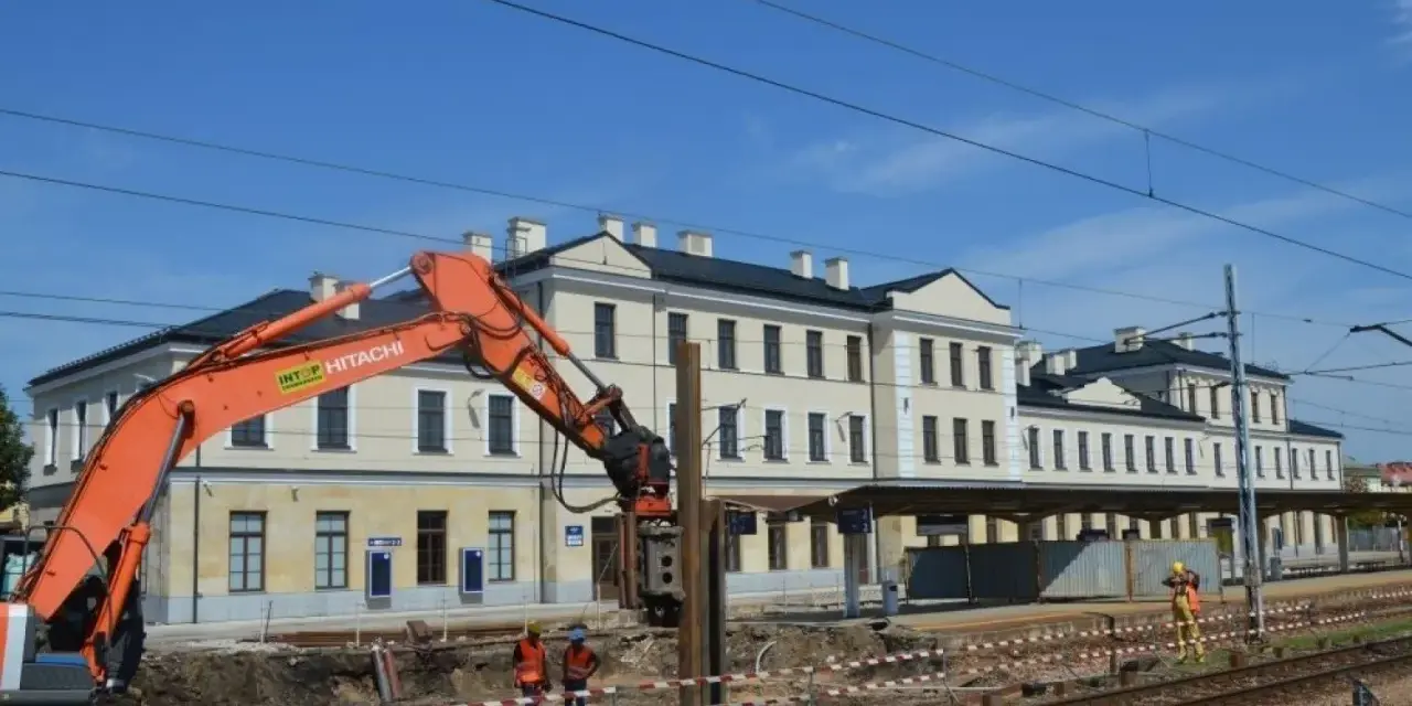 Koparka pracuje przy modernizacji dworca Skarżysko Kamienna. Widać budynek dworca, tory i pracowników w kamizelkach.