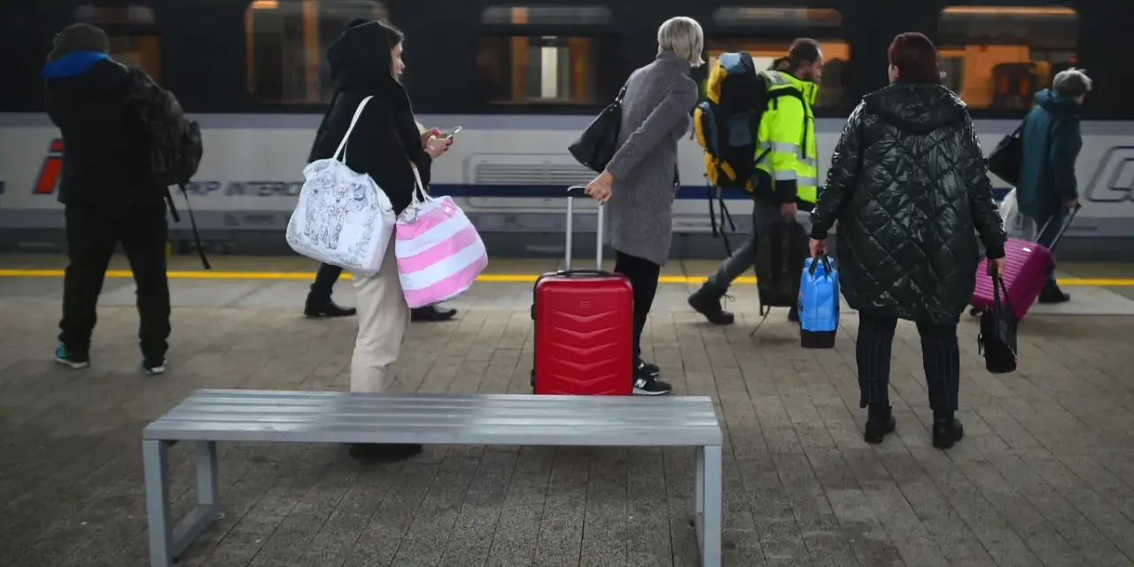 Ludzie z bagażami czekają na peronie PKP Intercity. Czerwona walizka stoi obok ławki.