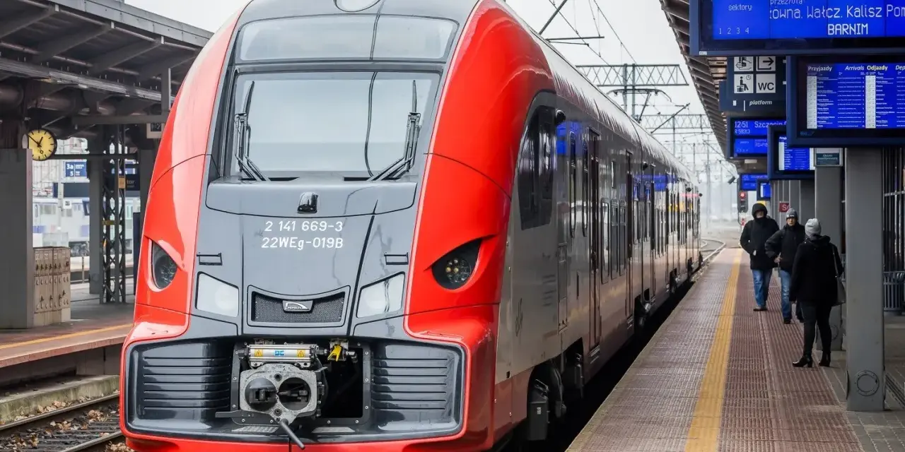 Czerwono-szary pociąg stoi na peronie, obok tablic informacyjnych z rozkładem jazdy.