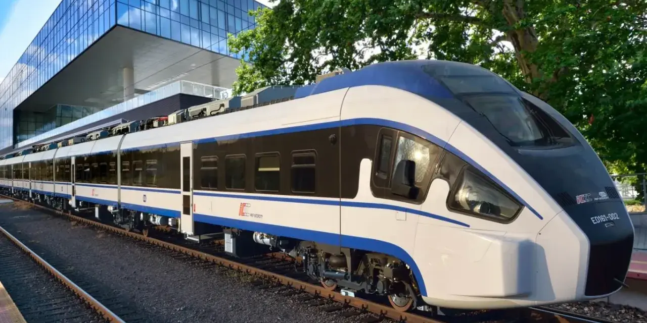 Nowoczesny, biało-niebieski pociąg Intercity ED161-002, najszybszy pociąg w Polsce, stoi na torach obok futurystycznego budynku.