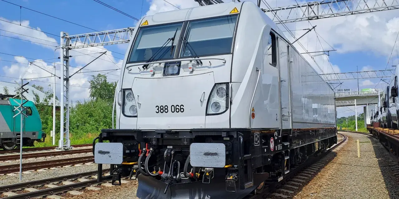 Nowy, biały lokomotywa 388 066, niczym nowoczesny bombardier, stoi na torach. W tle zielone drzewa i błękitne niebo.