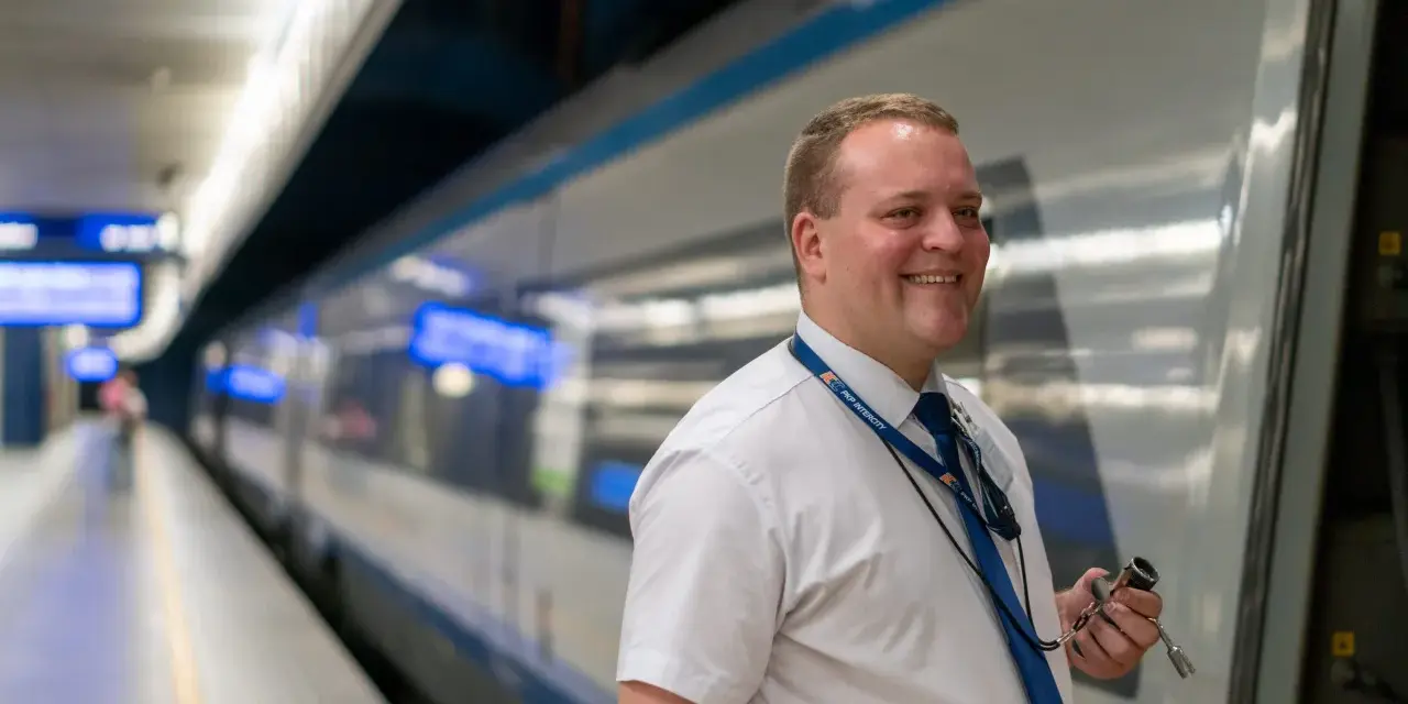 Uśmiechnięty konduktor PKP Intercity w białej koszuli i niebieskim krawacie stoi na peronie obok pociągu.