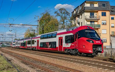 Nowoczesne, czerwono-białe piętrowe pociągi CFL 2410 pędzą po torach, mijając budynki i drzewa.