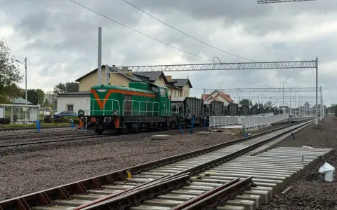 Zielona lokomotywa z pomarańczowymi pasami na torach. W tle budynki dworca i wagonów towarowych. Sprawdzam kętrzyn-olsztyn rozkład jazdy.