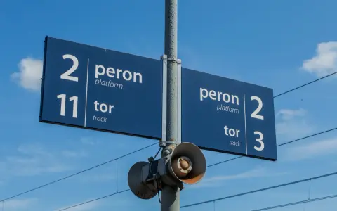 Niebieska tablica informacyjna na dworcu kolejowym wskazuje peron 2, tor 11 oraz peron 2, tor 3. W tle błękitne niebo z chmurami i linie trakcyjne.
