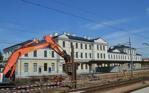 Koparka pracuje przy modernizacji dworca Skarżysko Kamienna. Widać budynek dworca, tory i pracowników w kamizelkach.