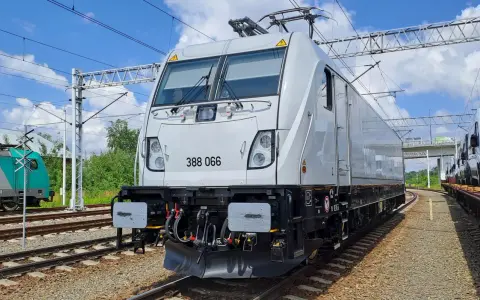 Nowy, biały lokomotywa 388 066, niczym nowoczesny bombardier, stoi na torach. W tle zielone drzewa i błękitne niebo.