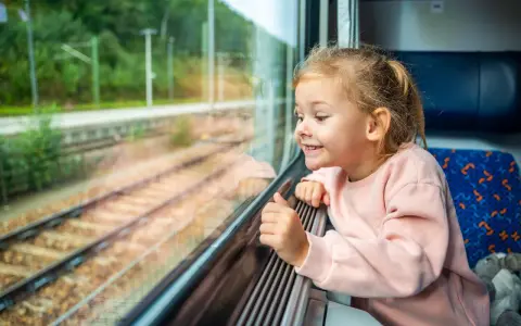Uśmiechnięta dziewczynka w różowym swetrze patrzy przez okno pociągu na tory.