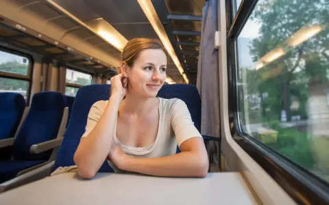 Uśmiechnięta kobieta w pociągu, patrząca przez okno. Czeka na kolejne przesiadki, ciesząc się podróżą.