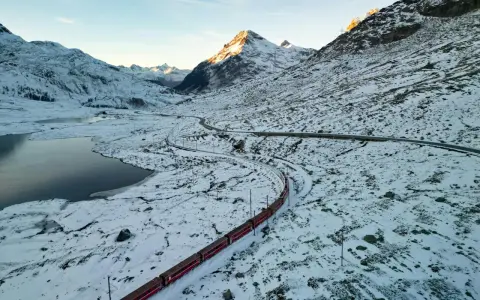 Czerwony pociąg panoramiczny wije się przez ośnieżony górski krajobraz. Słońce oświetla szczyty, a obok jezioro odbija błękitne niebo.