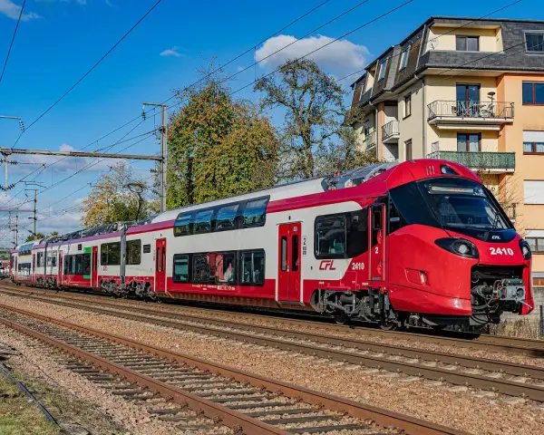 Nowoczesne, czerwono-białe piętrowe pociągi CFL 2410 pędzą po torach, mijając budynki i drzewa.