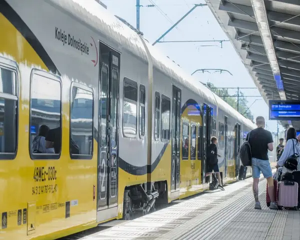 Pociąg Koleje Dolnośląskie na stacji. Ludzie z bagażami czekają na peronie, gotowi do podróży.