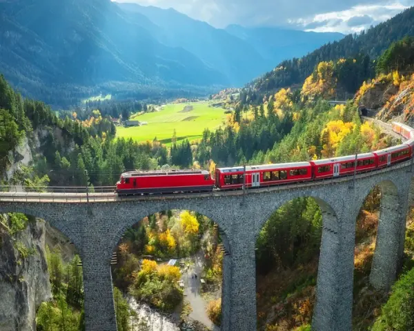 Czerwony pociąg Bernina Express przejeżdża przez kamienny wiadukt w górach jesienią.