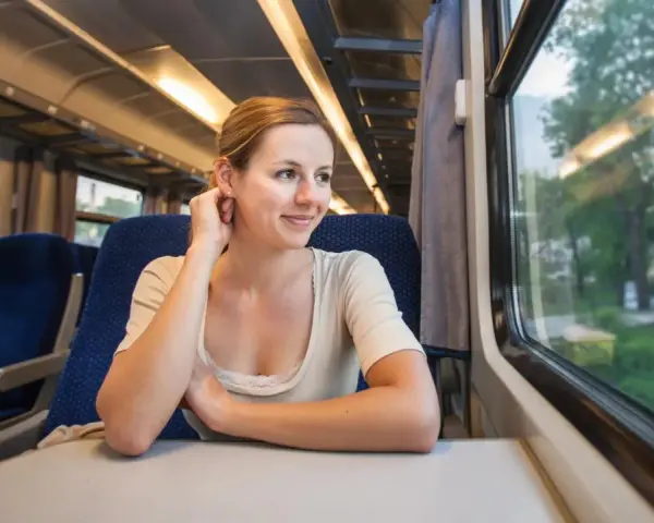 Uśmiechnięta kobieta w pociągu, patrząca przez okno. Marzy o podróży pociągiem do Włoch, widząc za szybą zielone krajobrazy.