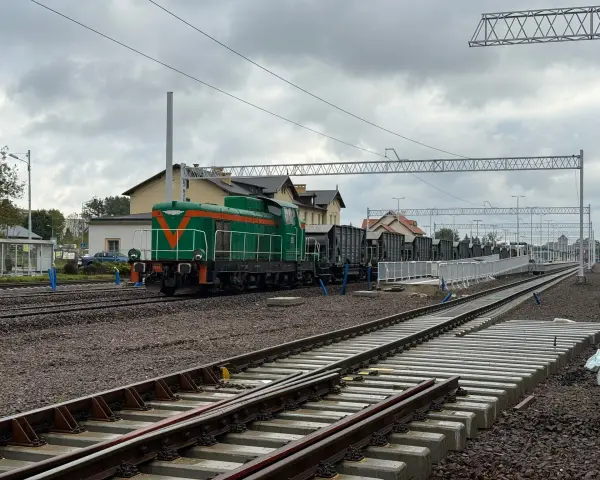 Zielona lokomotywa z pomarańczowymi pasami na torach. W tle budynki dworca i wagonów towarowych. Sprawdzam kętrzyn-olsztyn rozkład jazdy.