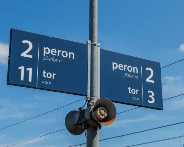 Niebieska tablica informacyjna na dworcu kolejowym wskazuje peron 2, tor 11 oraz peron 2, tor 3. W tle błękitne niebo z chmurami i linie trakcyjne.