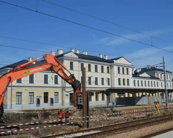 Koparka pracuje przy modernizacji dworca Skarżysko Kamienna. Widać budynek dworca, tory i pracowników w kamizelkach.