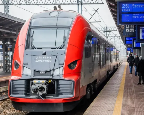 Czerwono-szary pociąg stoi na peronie, obok tablic informacyjnych z rozkładem jazdy.