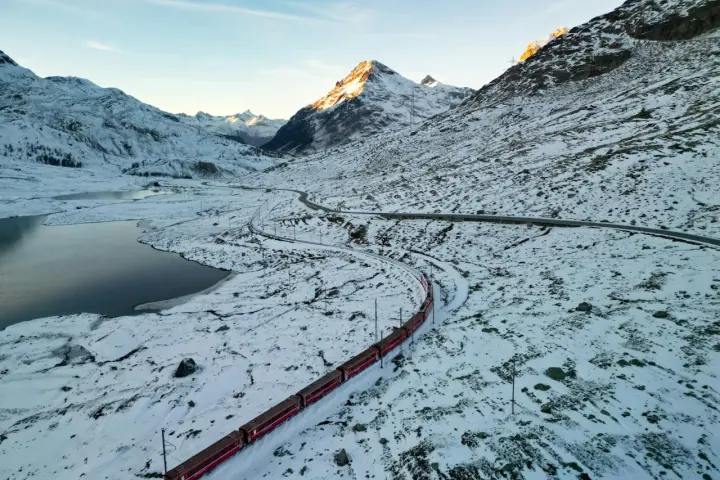 Czerwony pociąg panoramiczny wije się przez ośnieżony górski krajobraz. Słońce oświetla szczyty, a obok jezioro odbija błękitne niebo.