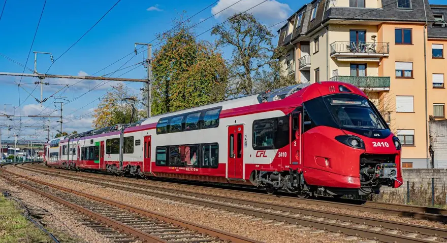 Nowoczesne, czerwono-białe piętrowe pociągi CFL 2410 pędzą po torach, mijając budynki i drzewa.