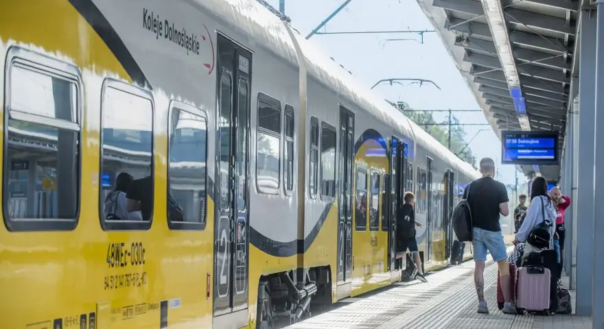 Pociąg Koleje Dolnośląskie na stacji. Ludzie z bagażami czekają na peronie, gotowi do podróży.
