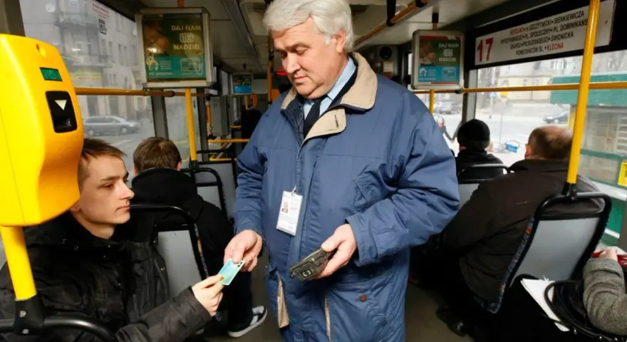 Kontroler biletów w niebieskiej kurtce sprawdza bilet młodego pasażera w autobusie.