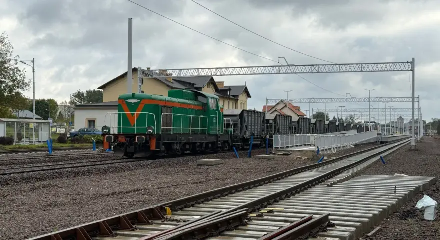 Zielona lokomotywa z pomarańczowymi pasami na torach. W tle budynki dworca i wagonów towarowych. Sprawdzam kętrzyn-olsztyn rozkład jazdy.