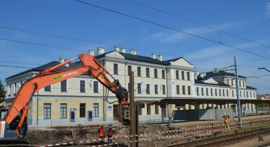 Koparka pracuje przy modernizacji dworca Skarżysko Kamienna. Widać budynek dworca, tory i pracowników w kamizelkach.