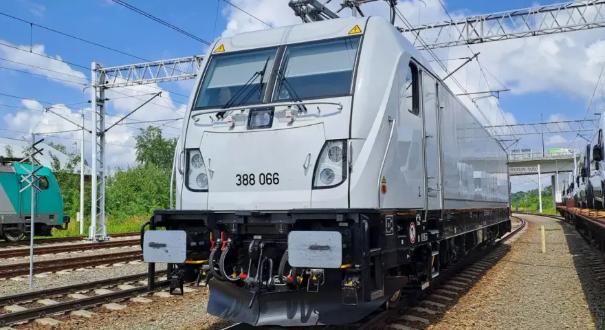 Nowy, biały lokomotywa 388 066, niczym nowoczesny bombardier, stoi na torach. W tle zielone drzewa i błękitne niebo.