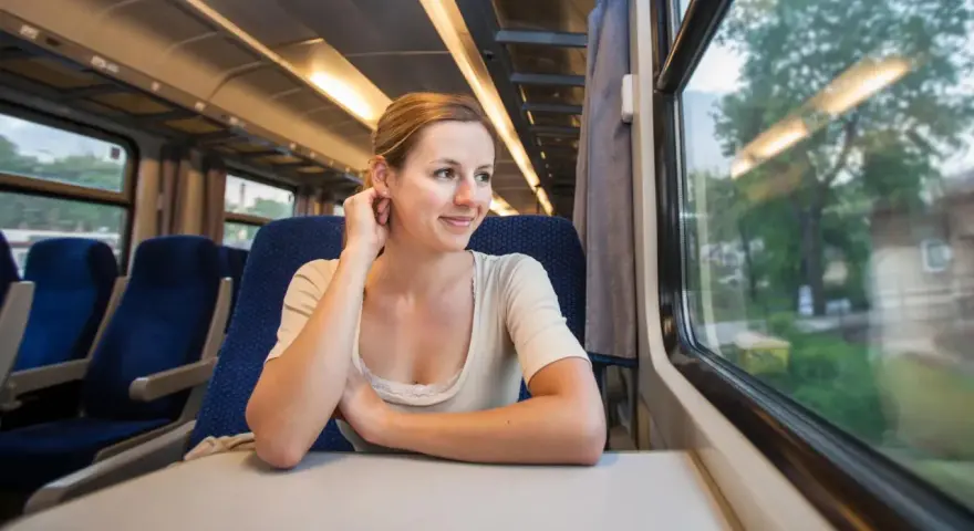 Uśmiechnięta kobieta podróżuje pociągiem, podziwiając krajobrazy, które mogą być częścią najpiękniejszych tras kolejowych w Europie.