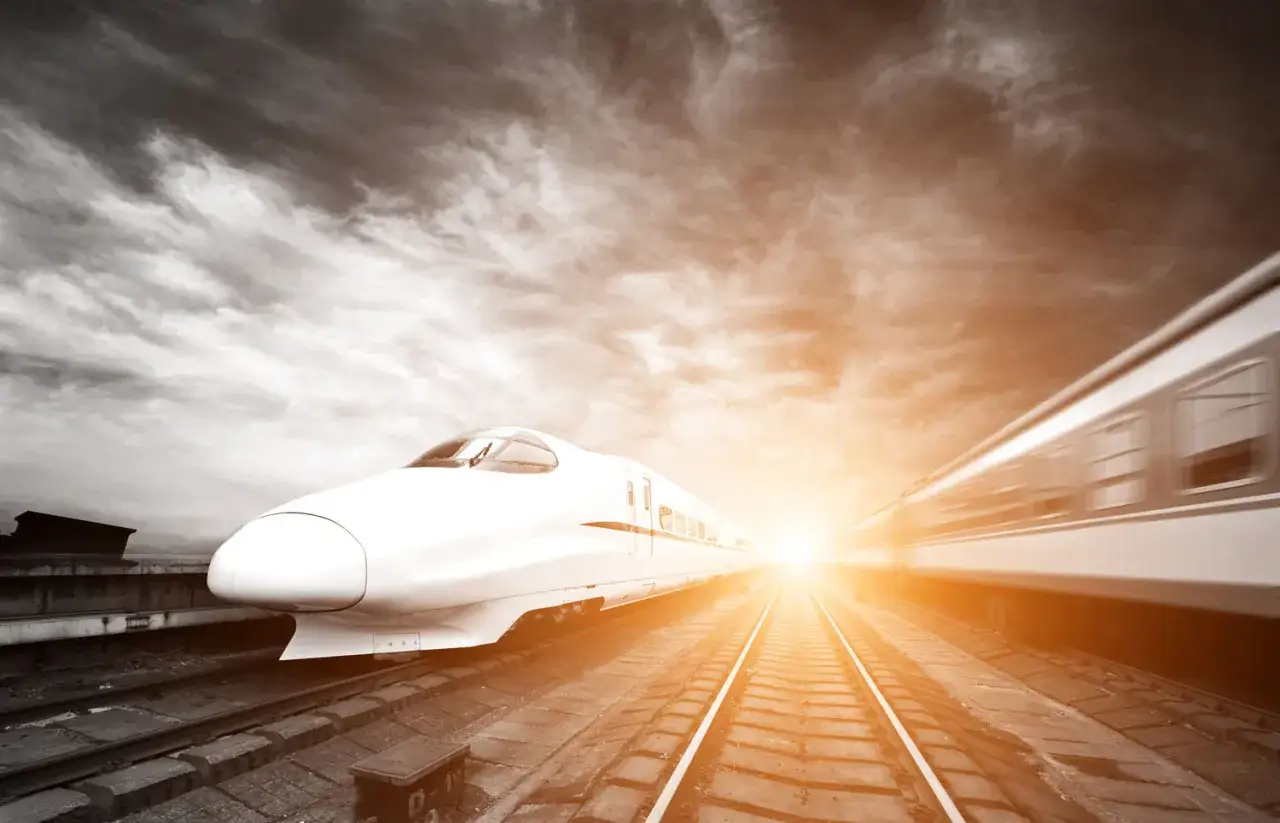 Biały shinkansen pędzi po torach w stronę zachodzącego słońca. W tle widać drugi pociąg w ruchu.
