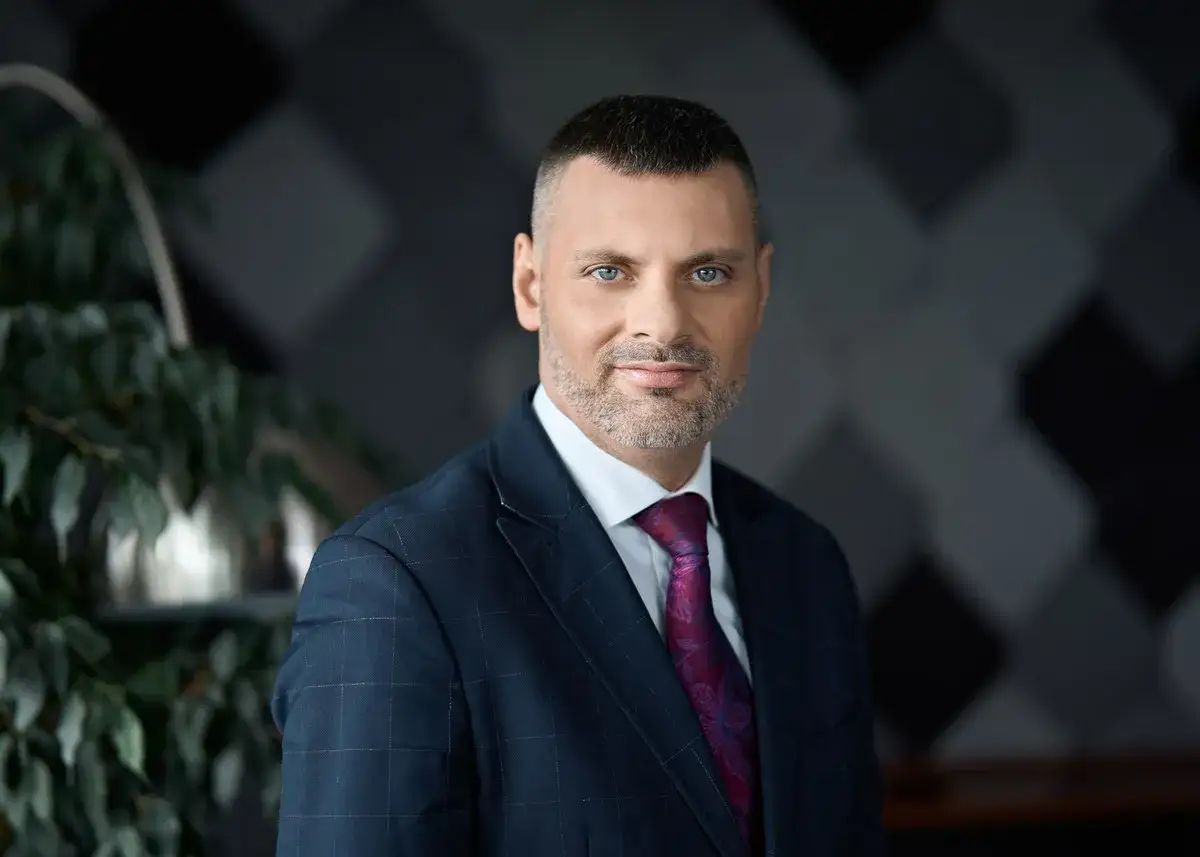 Prezes PKP w eleganckim garniturze, z niebieskimi oczami, patrzy wprost na widza. W tle roślinność i geometryczny wzór.