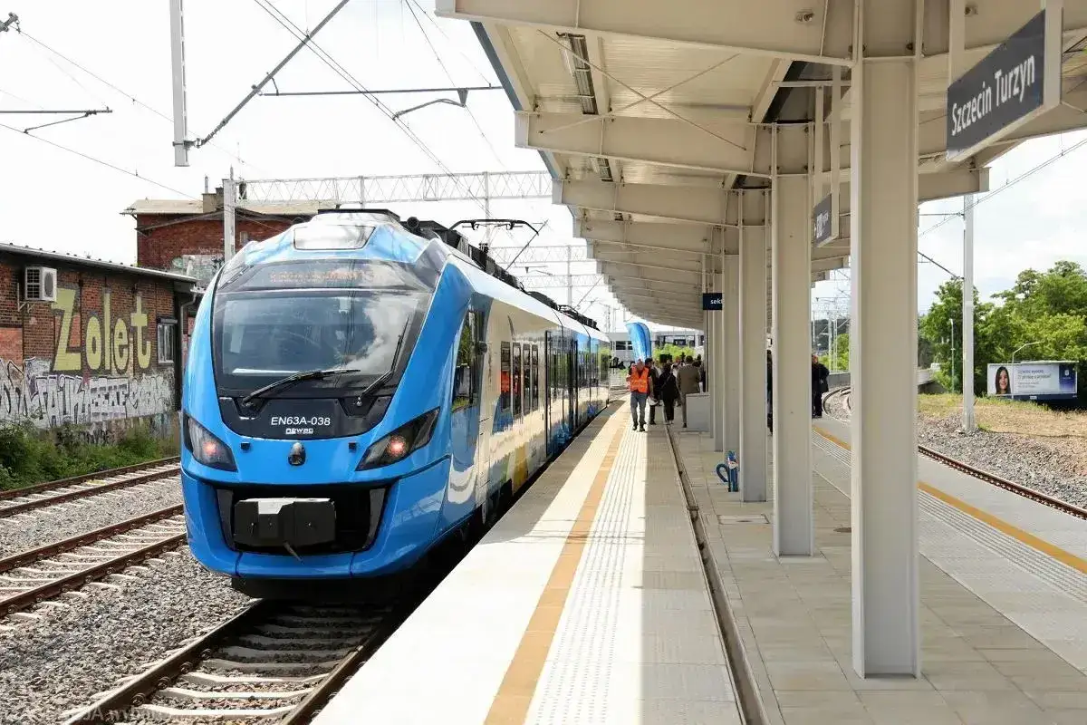Nowy, niebieski pociąg EN63A-038 na stacji Szczecin Turzyn. W tle budynek z graffiti i peron z pasażerami.