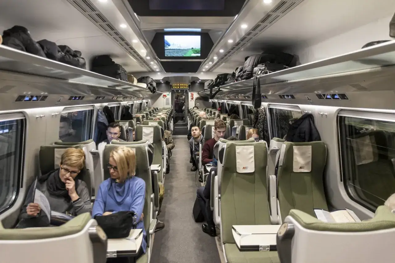 Wnętrze Pendolino, klasa 2. Pasażerowie siedzą w zielonych fotelach, bagaże na półkach. Komfort podróży w cenie biletu.