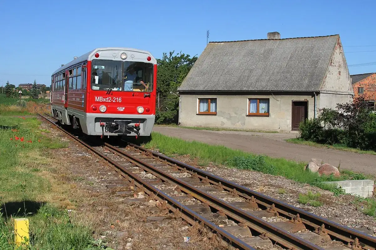 Czerwony pociąg SKPL MBxd2-216 jedzie po torach obok domu z szarym dachem.