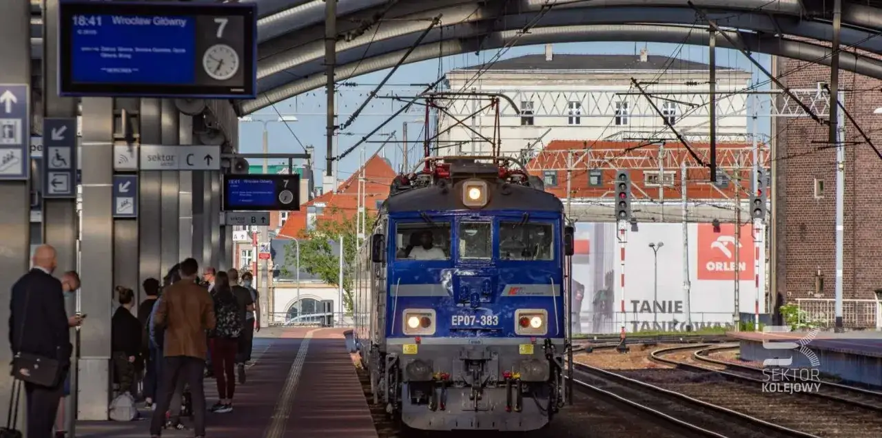 Niebieski pociąg PKP EP07-383 na stacji Wrocław Główny. W tle budynki i tablice informacyjne, gdzie można znaleźć informacje o PKP Biuro Rzeczy Znalezionych.