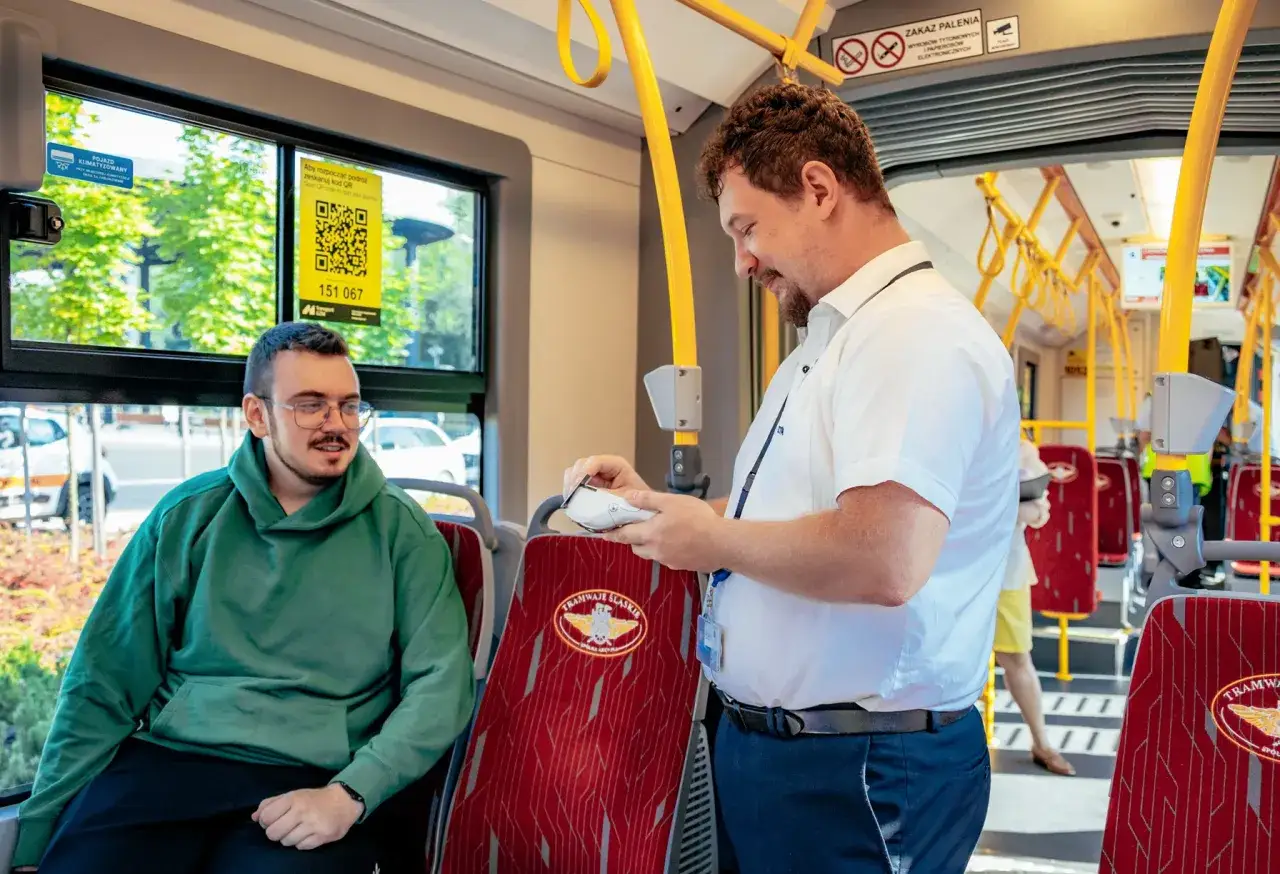 Konduktor w tramwaju sprawdza bilet. Młody pasażer w zielonej bluzie z kapturem czeka na kontrolę. Na siedzeniu widnieje logo 