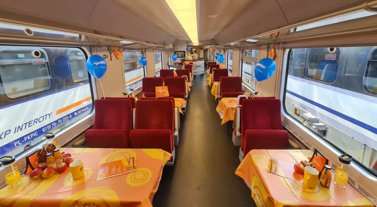 Wnętrze wagonu restauracyjnego PKP Intercity. Czerwone siedzenia, stoliki nakryte pomarańczowymi obrusami, balony i owoce tworzą przytulną atmosferę.