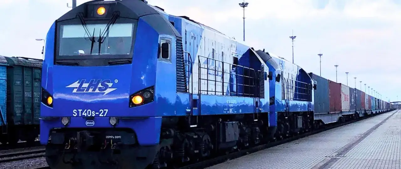 Niebieska lokomotywa LHS ST40s-27 z długim składem towarowym, gotowa do podróży z Hrubieszowa do Warszawy. Sprawdź rozkład jazdy!