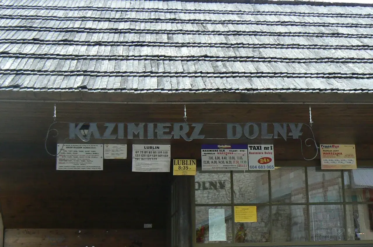 Tablica z rozkładem jazdy, informująca jak dojechać do Kazimierza Dolnego pociągiem i autobusem. Widoczne są nazwy miejscowości i godziny odjazdów.