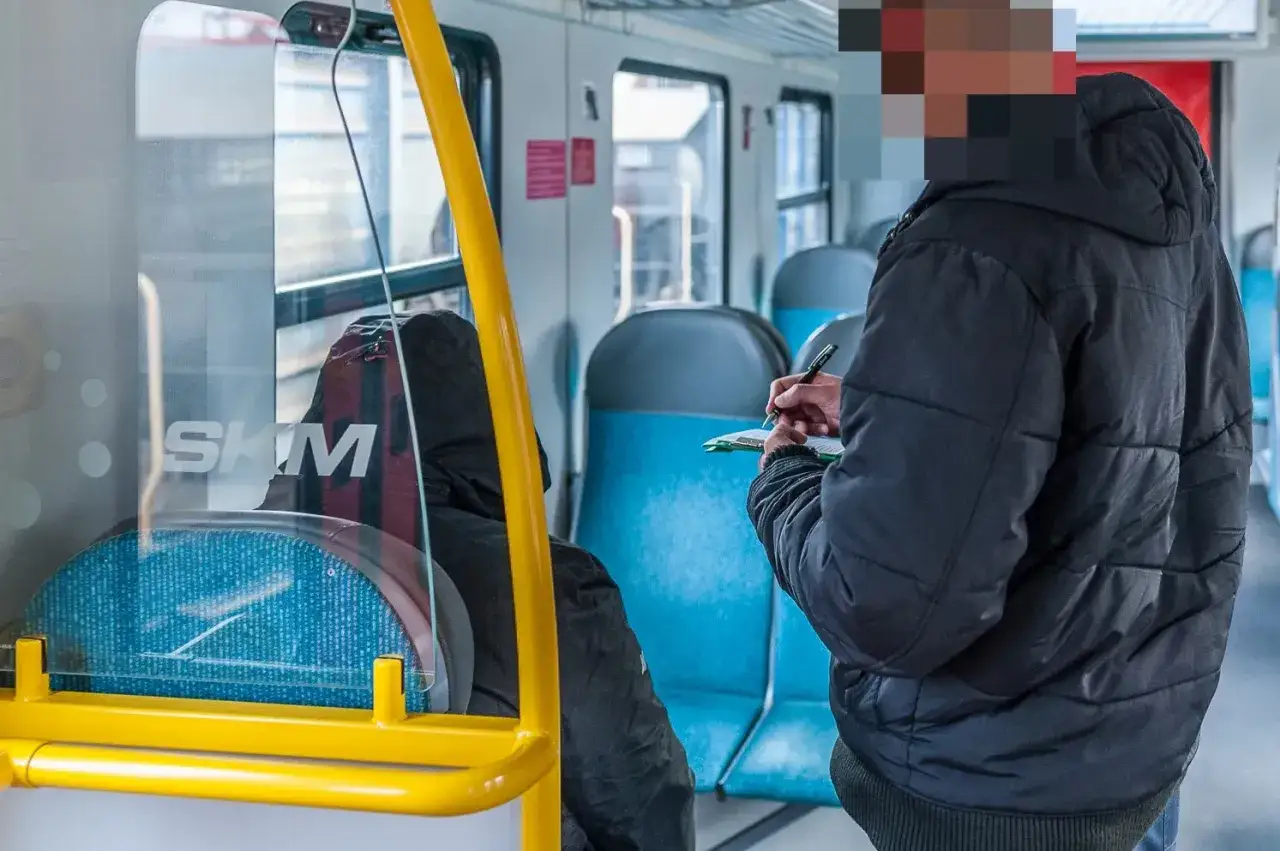 Konduktorka w SKM sprawdza bilety. W tle siedzą pasażerowie, a obok żółta poręcz.